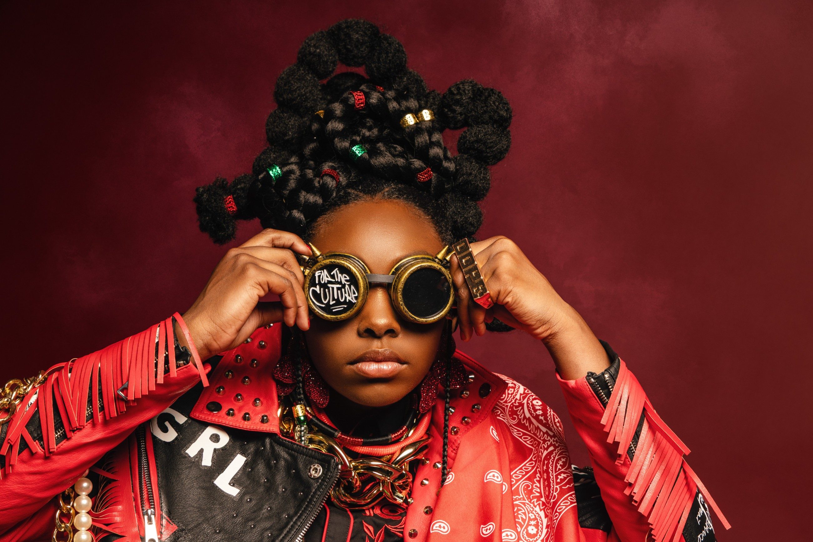 Girl in a red fringe jacket lifting goggles that read For The Culture