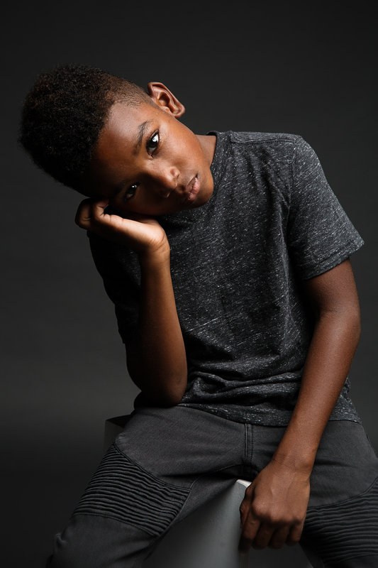 Boy in a grey tee resting a hand against his face on a dark backdrop
